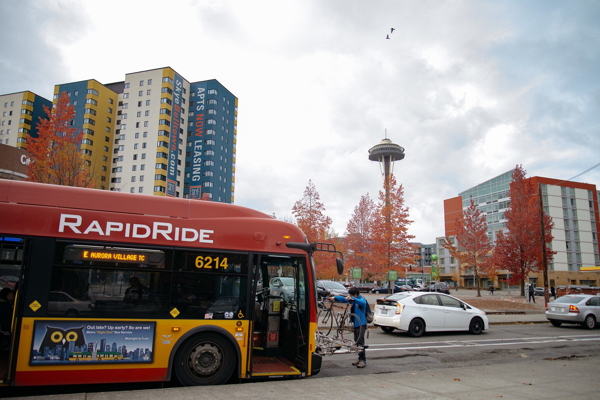 A person loads their bike and prepares to ride the northbound RapidRide E Line bus. The Space Needle stands tall in the background, alongside several large buildings.