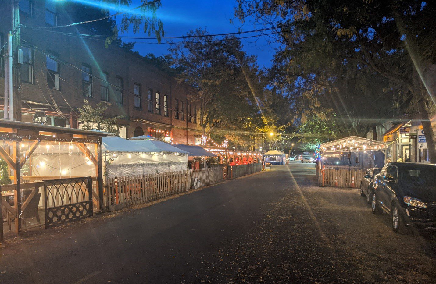 Several outdoor dining areas line the street on Ballard Ave NW in the evening. Large trees and buildings are visible in the background, along with parked cars on the right side.