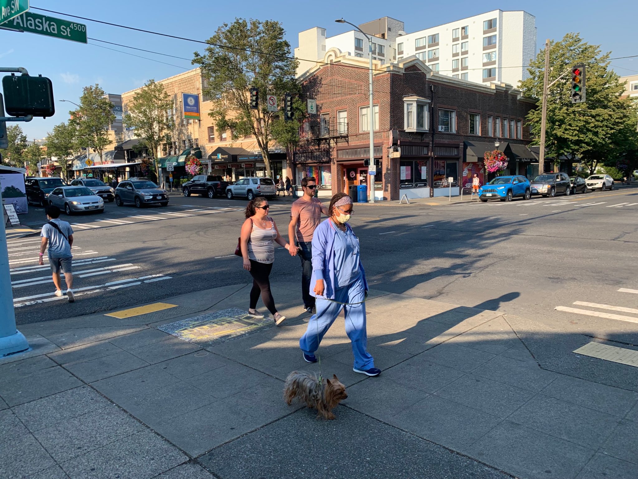 People walk through West Seattle along SW Alaska St. A woman wearing medical scrubs walks her dog while two people hold hands and a person walks across the street on the left side. Cars and buildings are visible in the background.