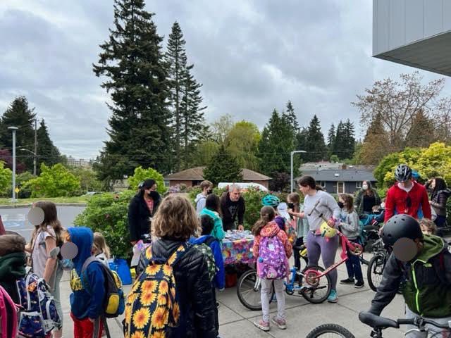 Students gather at the entrance to Hazel Wolf K-12 school after biking to school as part of their Bike to School event. Many students and adults are visible on the cloudy day at the entrance to the school.