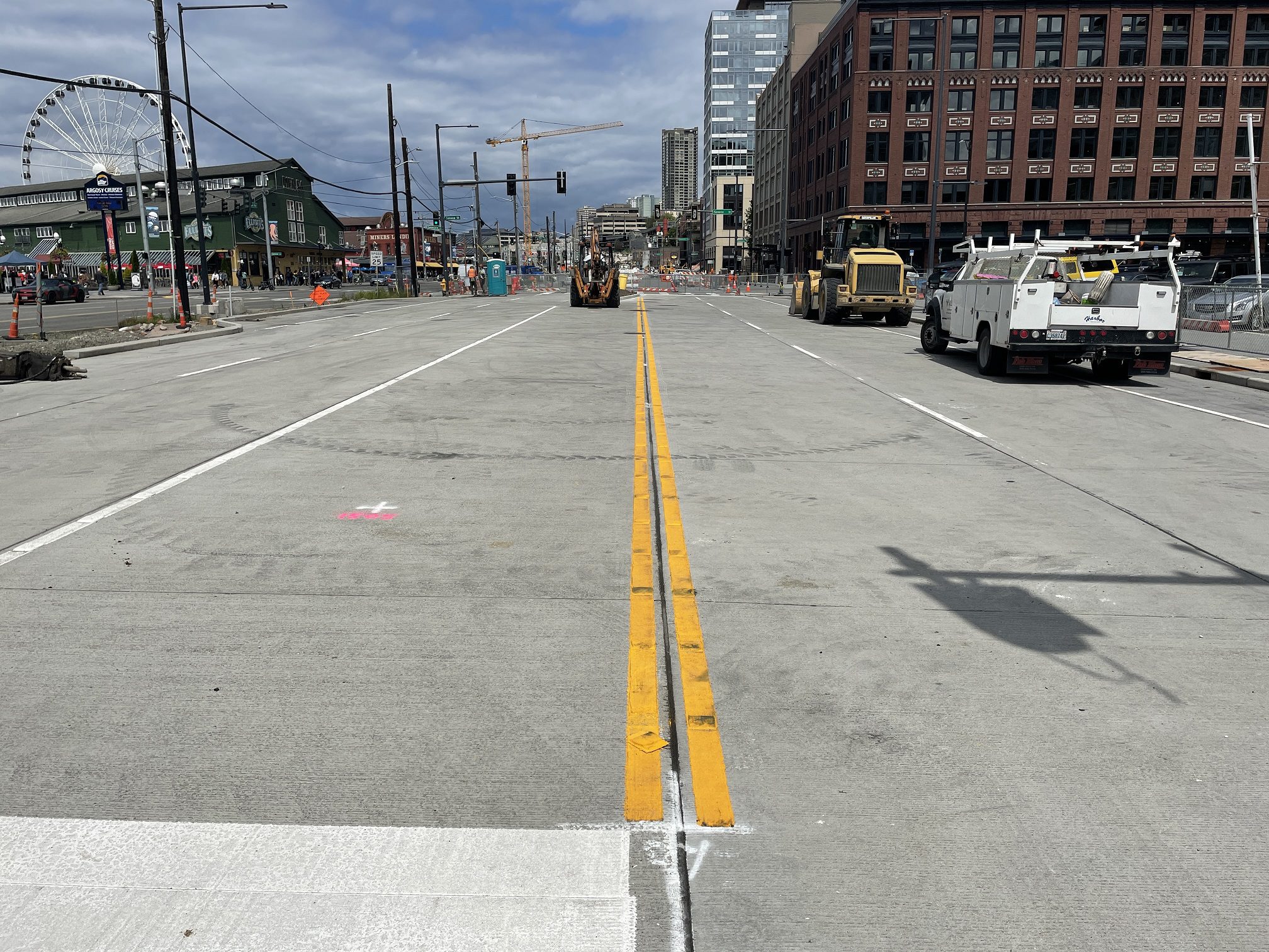 Lane markings on new travel lanes between Spring and Seneca streets in preparation to open five blocks of the newly constructed Alaskan Way. A double yellow line runs down the middle of the image, with work vehicles and large buildings in the background.