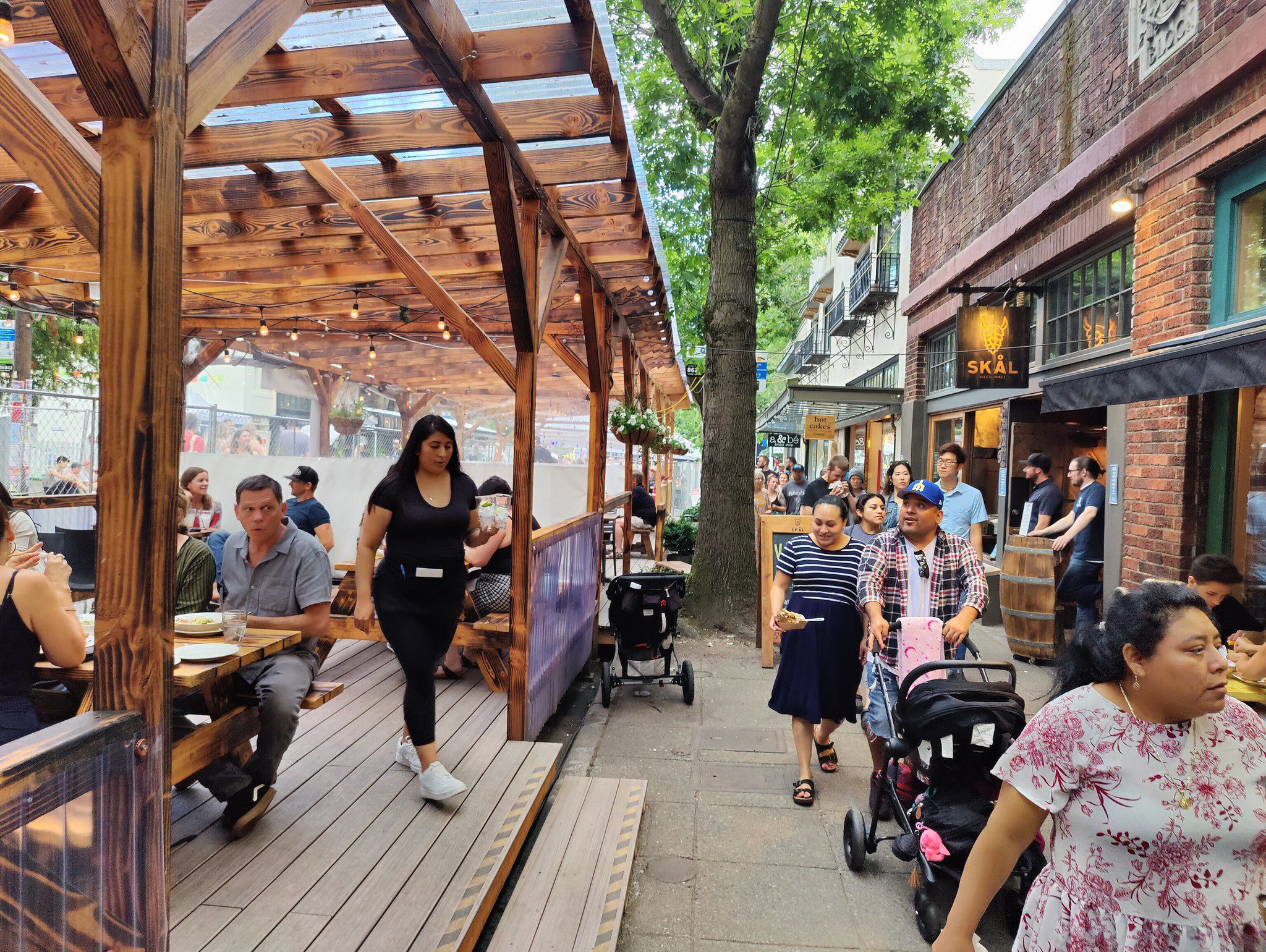 People enjoying an outdoor meal and walking on the sidewalk along Ballard Ave NW, on a busy summer weekend. Many people are visible sitting and walking, with a large tree in the middle and buildings on the right.