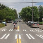 A woman crosses the street at 17th Ave SW and SW Henderson St. A marked crosswalk and rectangular rapid flashing beacon help make drivers aware of people crossing the street. Many mature trees and parked cars are present in the background and middle of the photo.