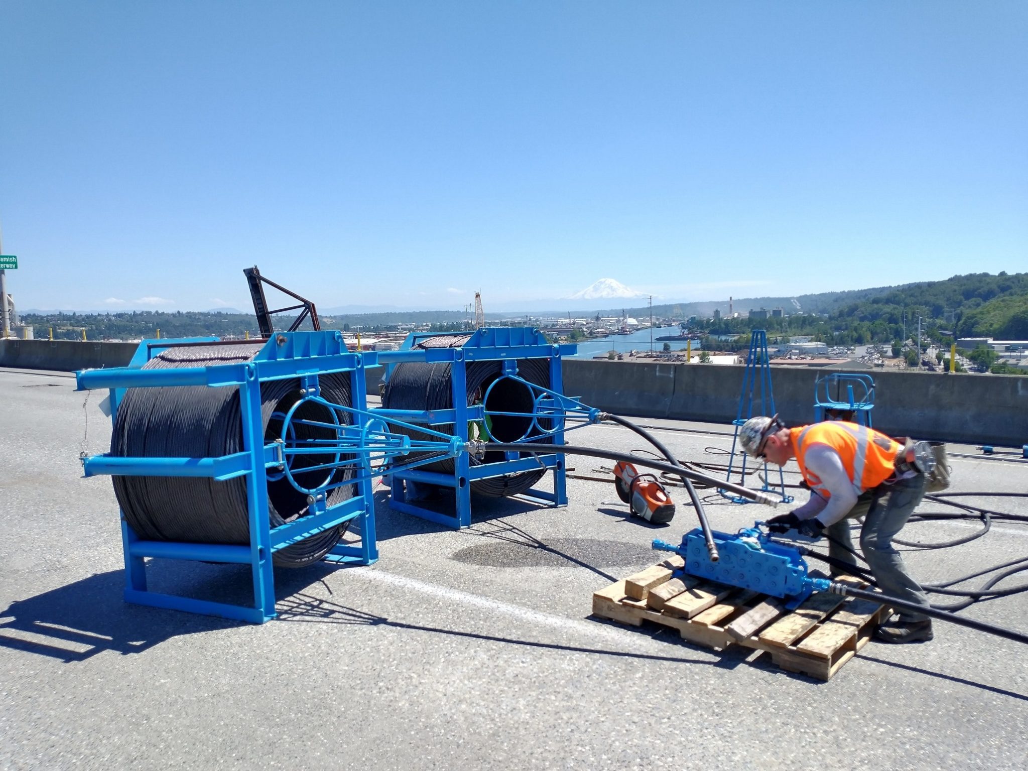 Steel cable is installed through protective pipes as part of the West Seattle Bridge’s newest post-tensioning system. A worker wearing an orange safety vest and hardhat works on the cables atop the bridge. Mount Rainier and the Duwamish Waterway are in the background.