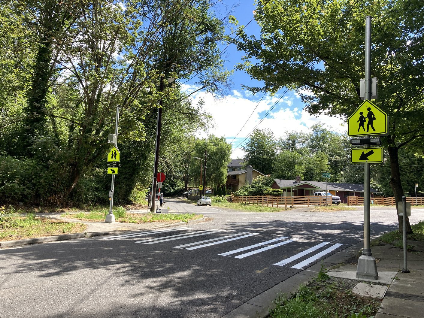 A newly-installed rectangular rapid flashing beacon at a crosswalk at Dumar Way SW and SW Orchard St. Mature trees, homes, and parked cars are present in the background.