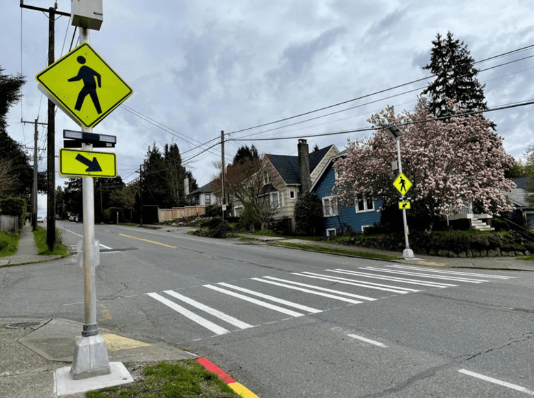 A marked crosswalk with rapid flashing beacons to alert drivers when pedestrians are crossing the street. The street and crosswalk take up most of the middle of the photo, with houses and trees in the background.