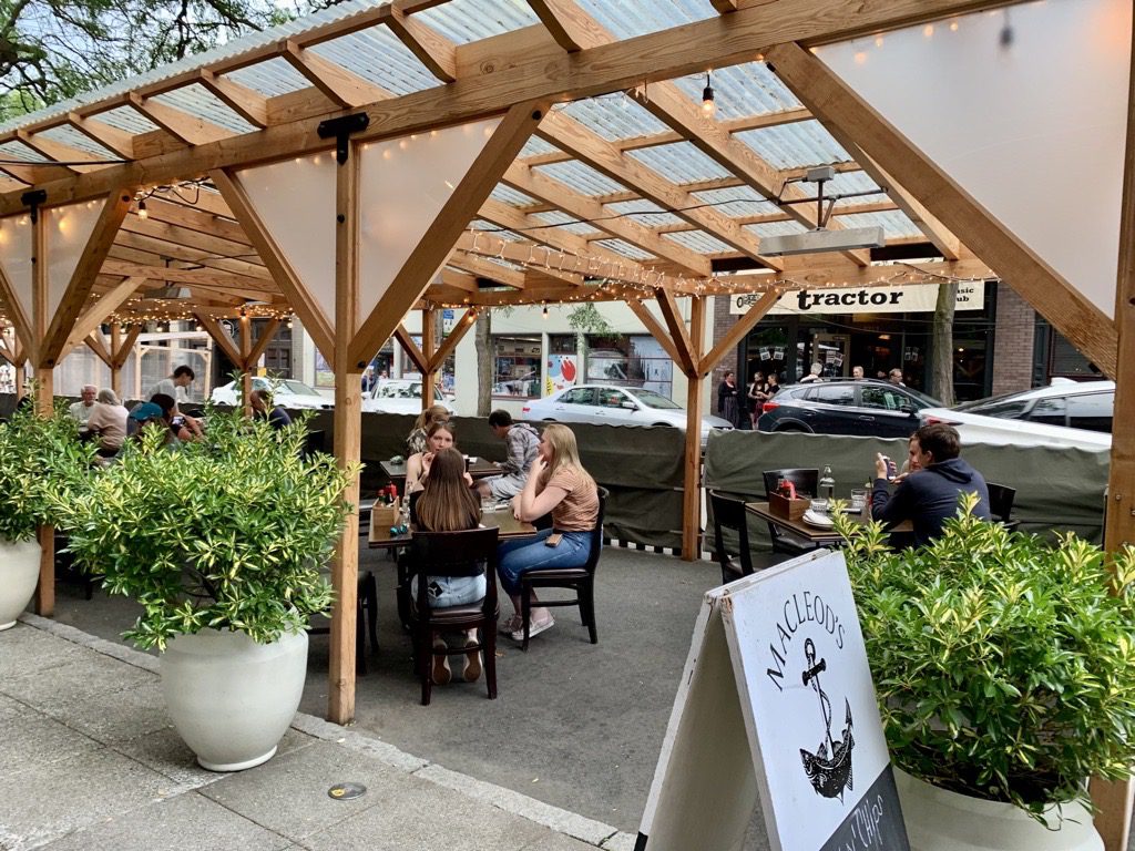 People enjoying a meal outdoors at a restaurant along Ballard Ave NW. Potted plants and other buildings are in the foreground and background.