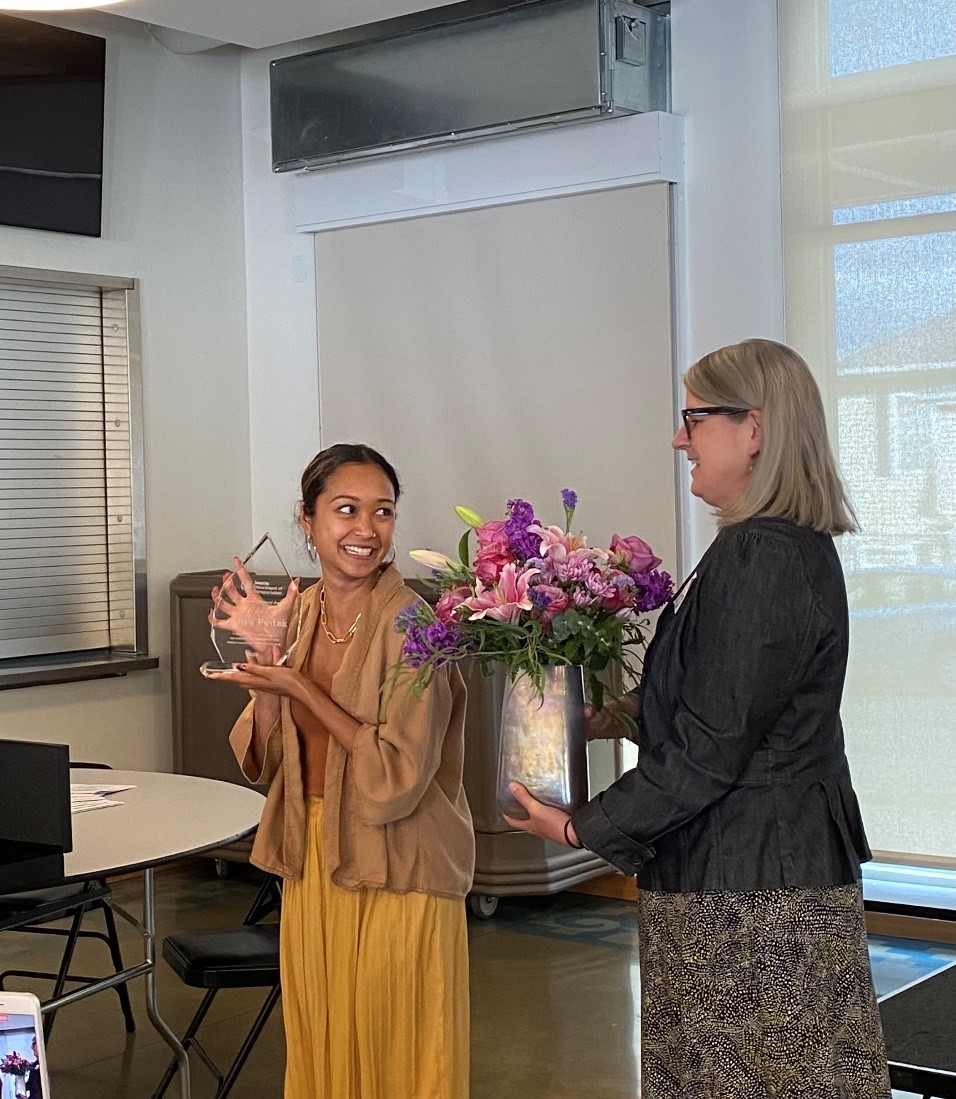 SDOT colleague holding a bouquet of pink and purple flowers in a metal vase while another colleague holds clear glass award smiling. 