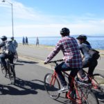 Several people bike in West Seattle on a sunny day. People walk in the background, near the water.