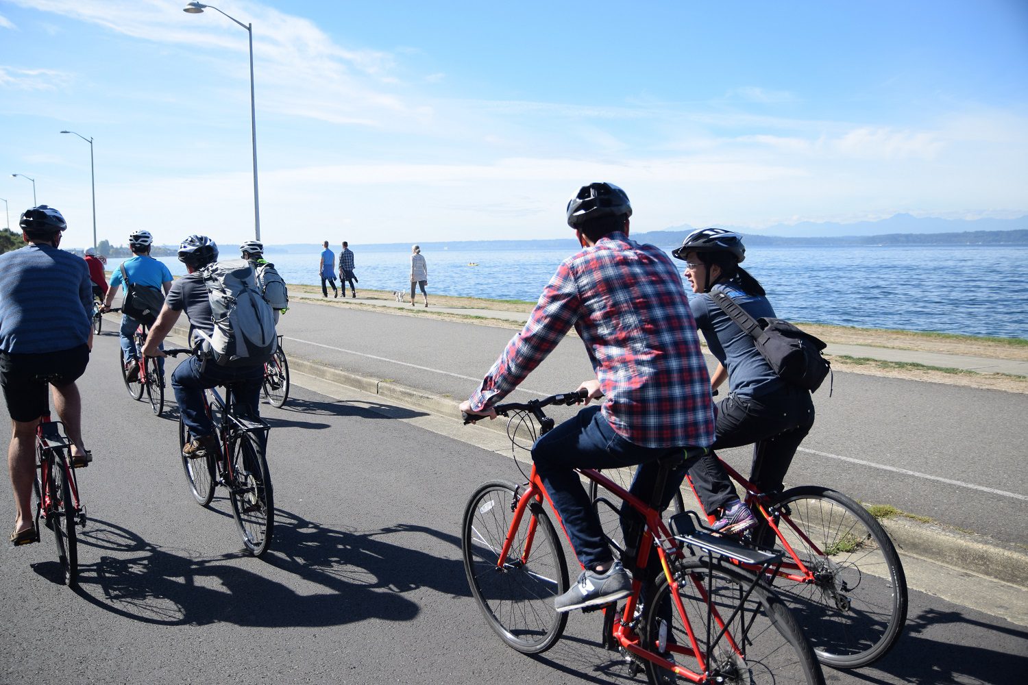 Several people bike in West Seattle on a sunny day. People walk in the background, near the water.