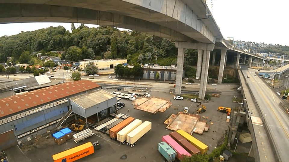 The West Seattle Bridge and two work platforms being lowered to the ground. Trees and construction equipment are also present in the background and foreground.