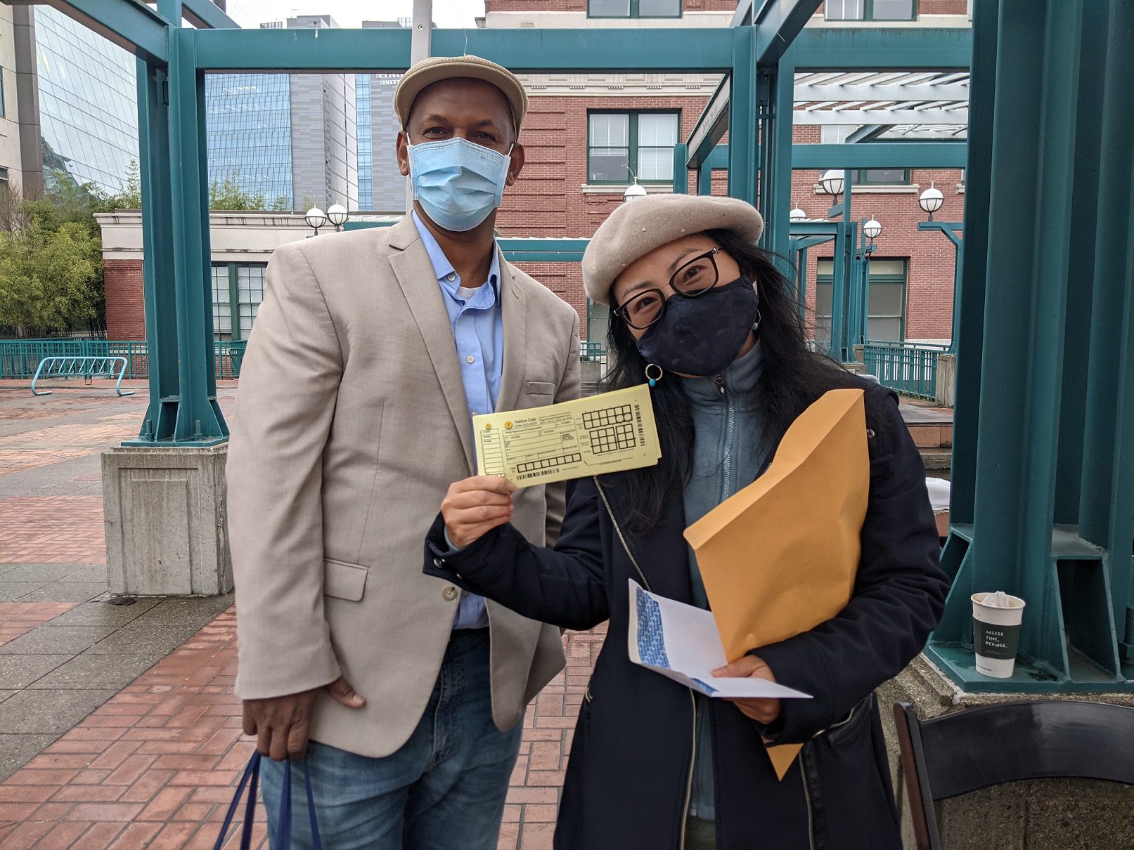 Two people standing outside wearing masks smile at the camera. The women on the right side holds up paper vouchers from the Ride Now pilot program. Large brick buildings are present in the background.