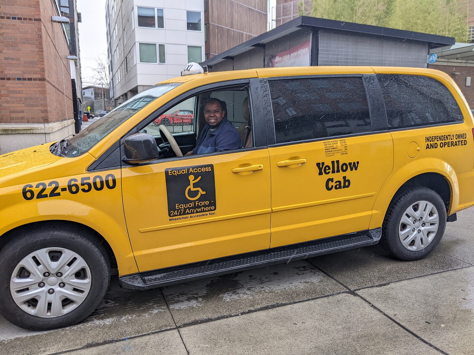 A Yellow Cab driver smiles at the camera on a rainy and windy day, while he waits to pick up a Ride Now pilot program participant in Seattle. Large buildings are present in the background.