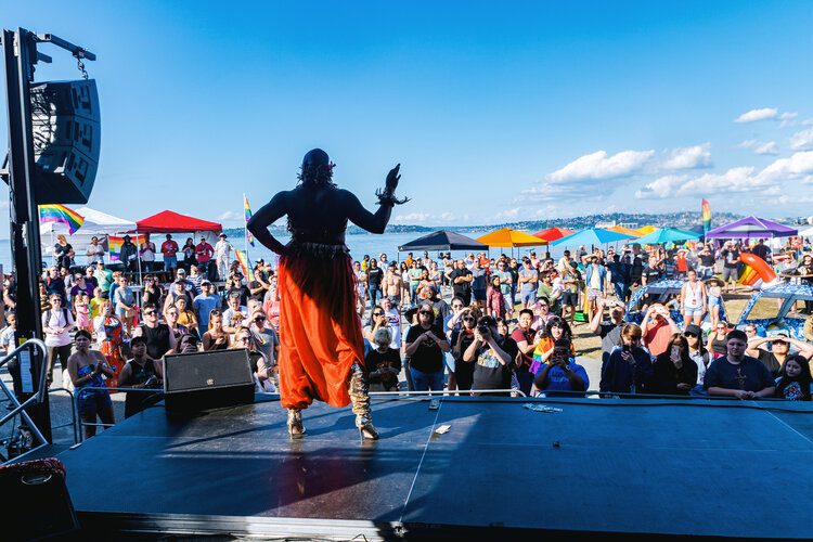 A performer on stage at a past Alki Beach Pride event, wearing a bright orange dress, in front of a large crowd on a sunny day.
