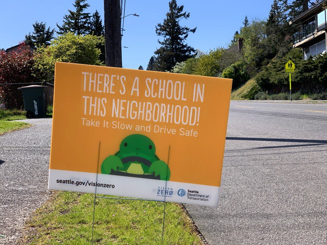 Orange yard sign with text "There's a school in this neighborhood! Take it slow and drive safe" in white letting with a graphic of a green turtle holding on to a steering wheel.