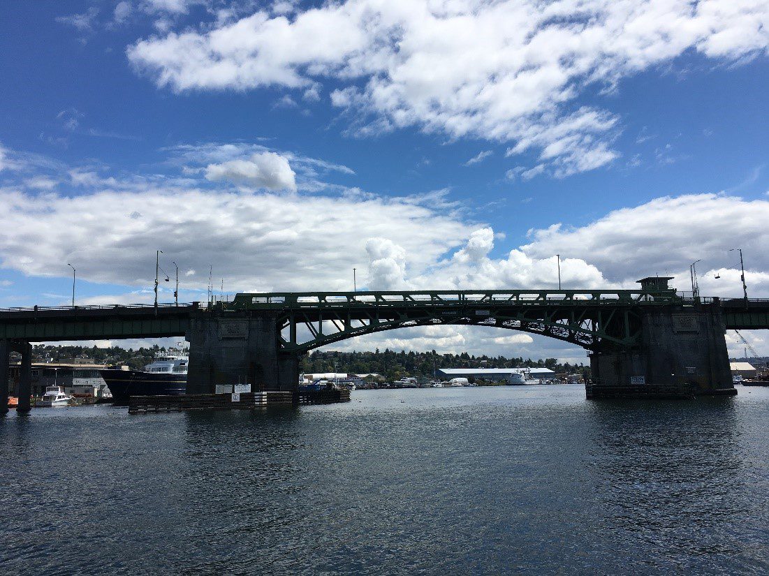 Photo of the Ballard Bridge from the side, with Lake Union in front and a cloudy sky.  
