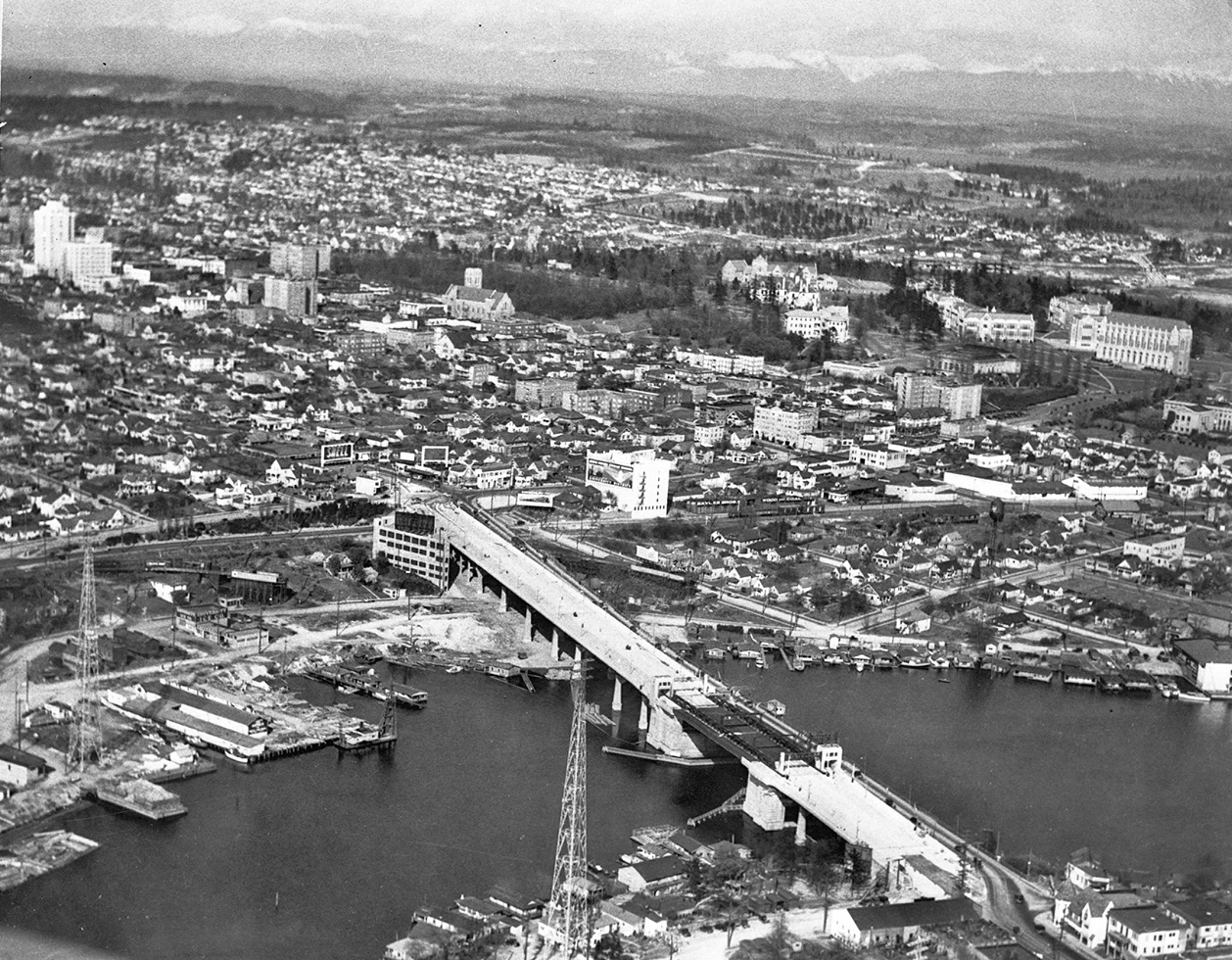 Black and white photo of the university bridge taken in an aerial shot with the University District behind it. 