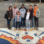 Dahvee Enciso with his family and father Javier at the ribbon cutting ceremony of the Dia De Los Muertos crosswalk dedicated to his mother Eva. The group stands on a mural in the street, smiling at the camera.