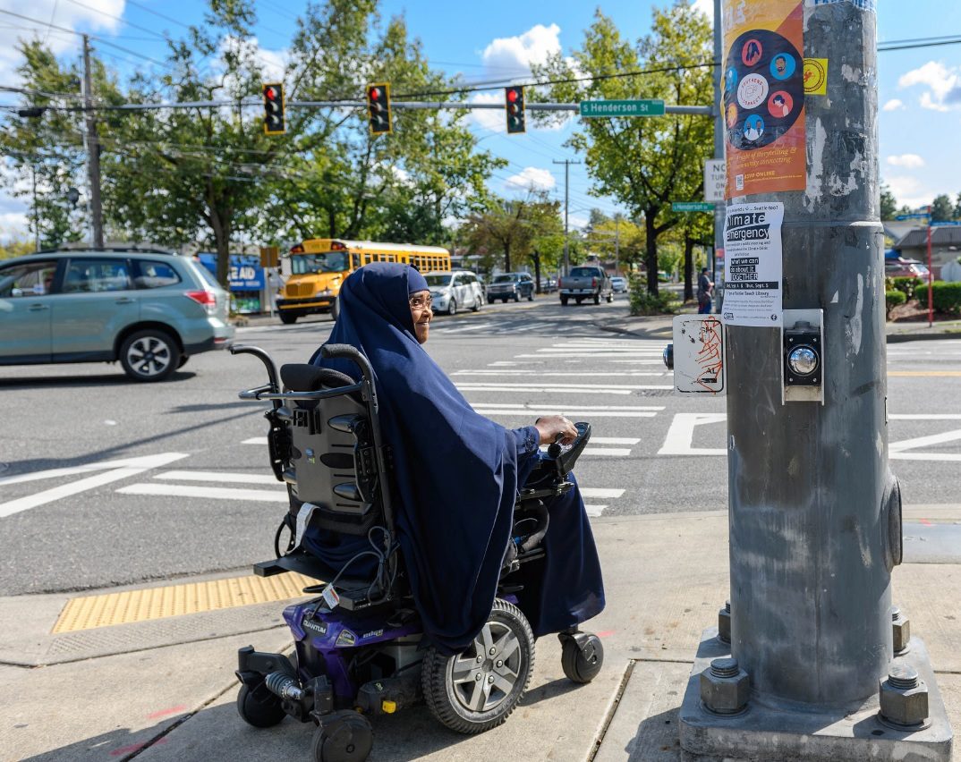 A woman using a power wheelchair prepares to cross the street in Southeast Seattle on a sunny day. Cars and a yellow school bus travel on the street in the background, and several mature trees stand in the background.