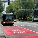 A recently constructed bus-only lane on Rainier Ave S. The bus travels in a lane painted red with the words "Bus Only" in white lettering. Large trees and traffic signals are in the background.