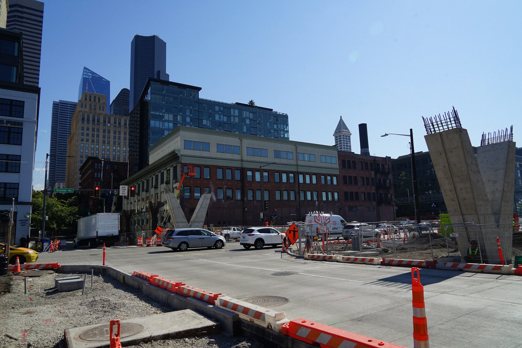Marion St pedestrian bridge construction crossing Alaskan Way. Large buildings and clear sky are present in the middle and top of the photo, with vehicles in the middle.