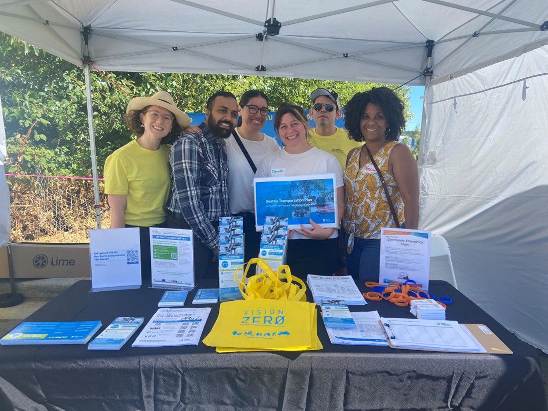 Photo of six department of transportation and neighborhood staff in yellow, plaid, and white shirts smiling behind an SDOT booth filled with flyers, pamphlets, and SDOT swag such as yellow Vision Zero totes and multi-colored bracelets.
