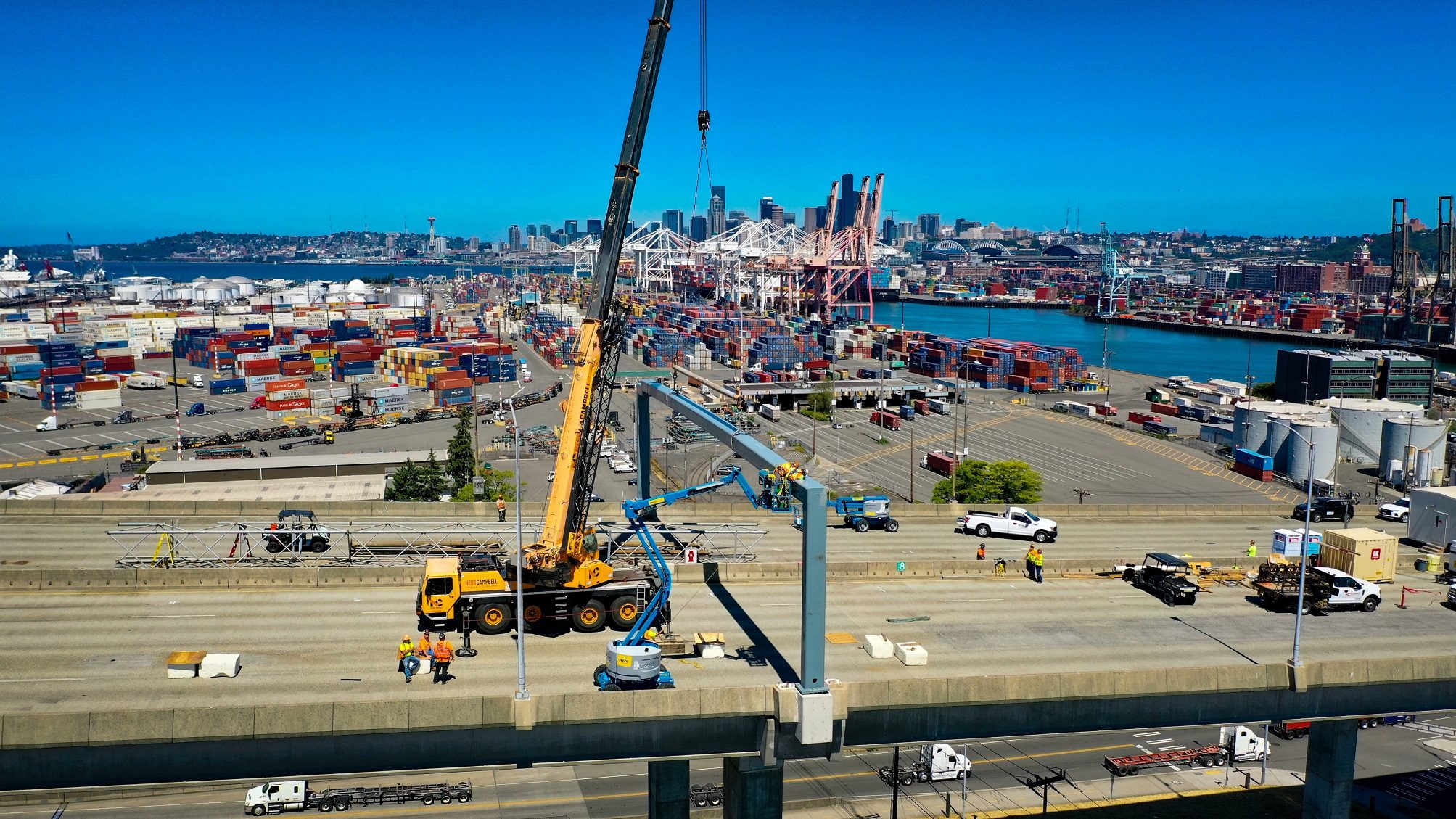Photo of a large crane on the West Seattle Bridge installing a sign structure. The Port of Seattle and downtown Seattle buildings are in the background on a sunny day.