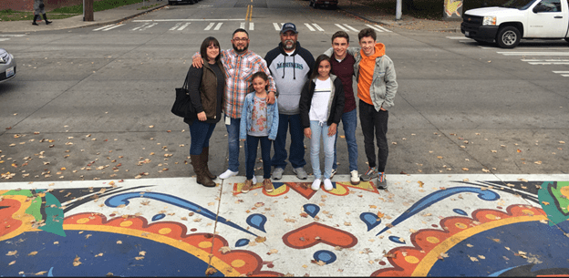 Dahvee Enciso with his family and father Javier at the ribbon cutting ceremony of the Dia De Los Muertos crosswalk dedicated to his mother Eva. The group stands on a mural in the street, smiling at the camera.