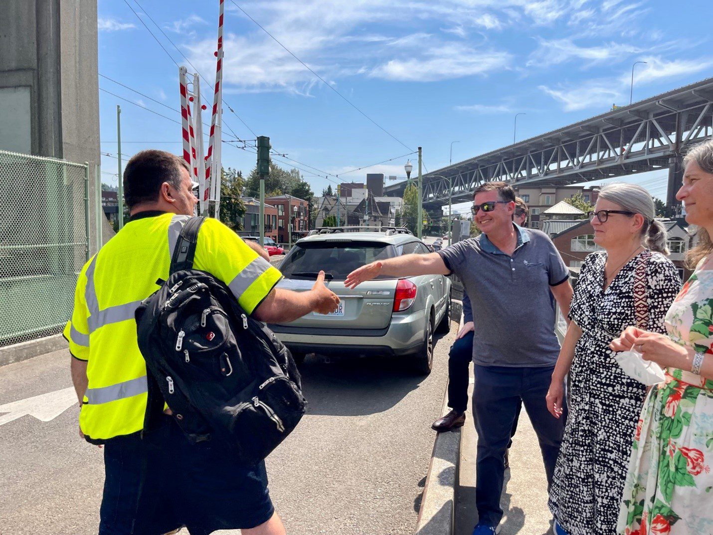 Greg Spotts shakes hands with SDOT team member Jared Hickman while touring the University Bridge in Seattle on a sunny day. A car and the Ship Canal Bridge are in the background.