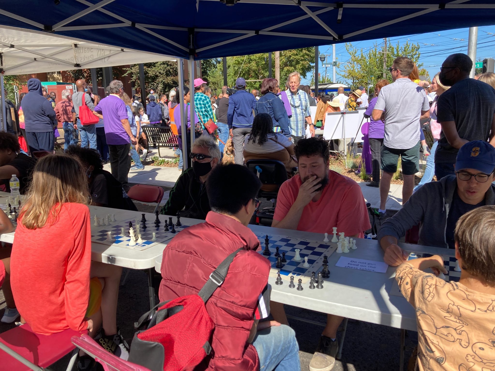 Kids and adults play the first games of chess at the newly opened Detective Cookie Chess Park in Rainier Beach. A row people playing chess at a table is in the foreground, with a crowd of people attending the celebration standing in the background.