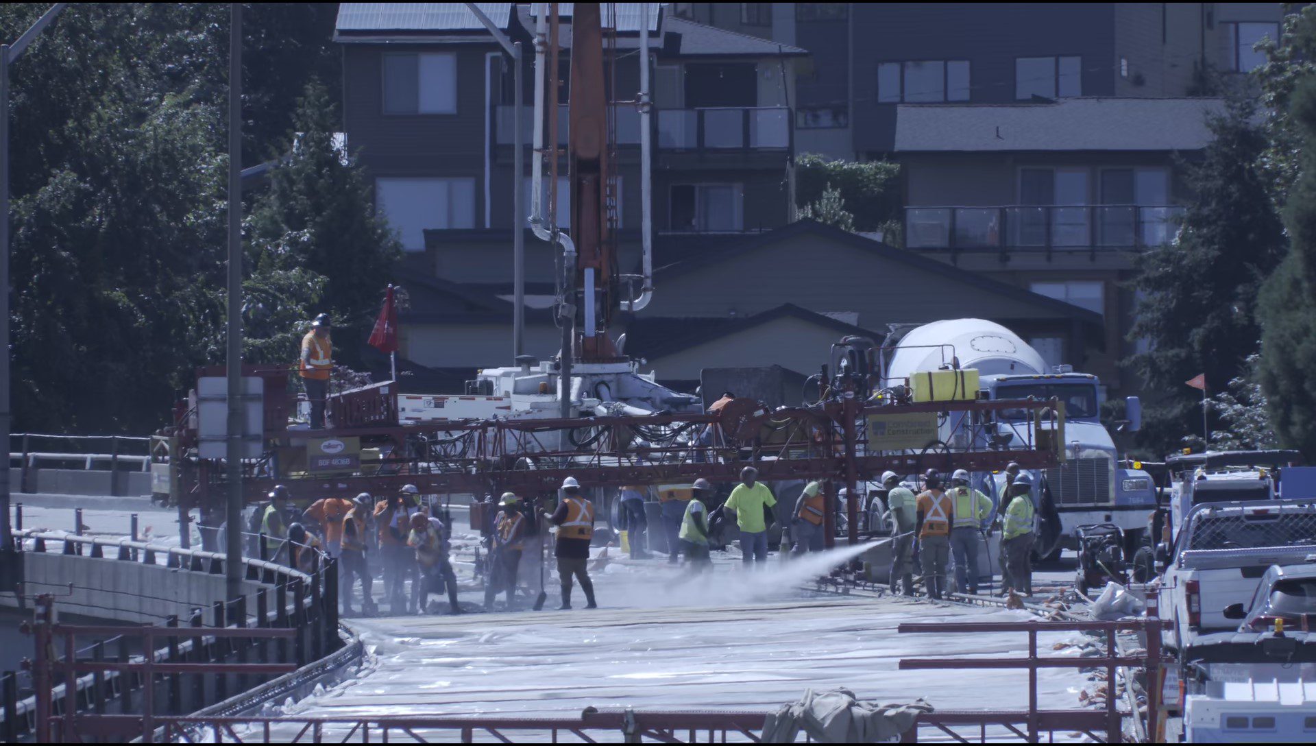 Numerous construction workers on the Fauntleroy Expressway, as part of a concrete overlay effort. Large construction equipment and machinery is in the center of the photo, with a large building in the background.