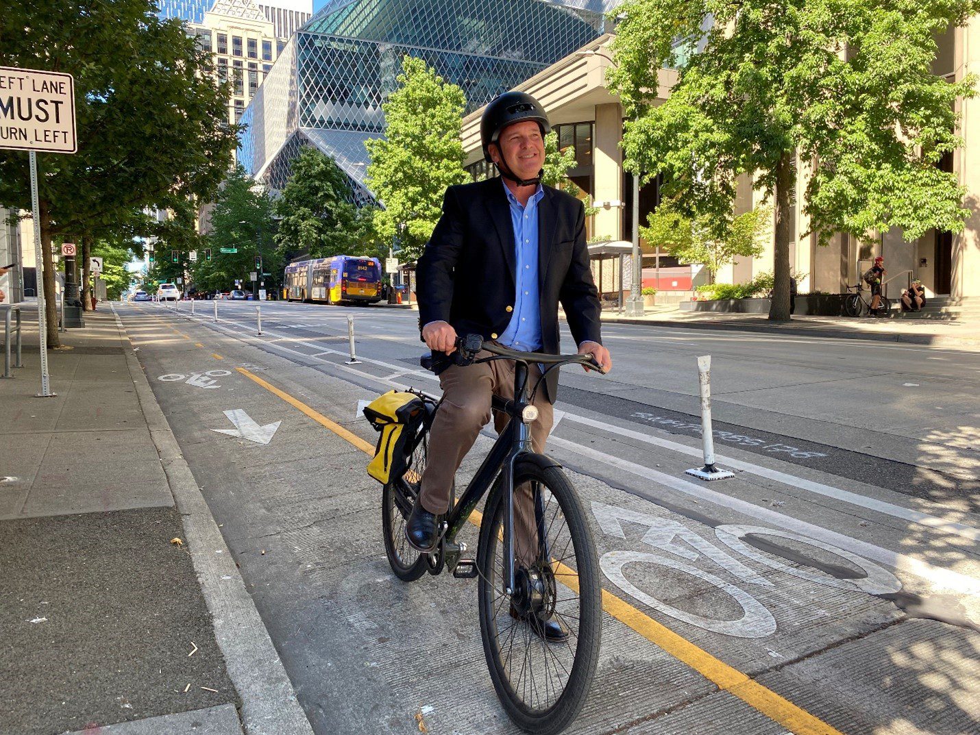 Greg Spotts bikes down the 4th Ave Protected Bike Lane in downtown Seattle on a sunny day. The Seattle Central Library and a King County Metro bus are in the background, as well as several large trees.