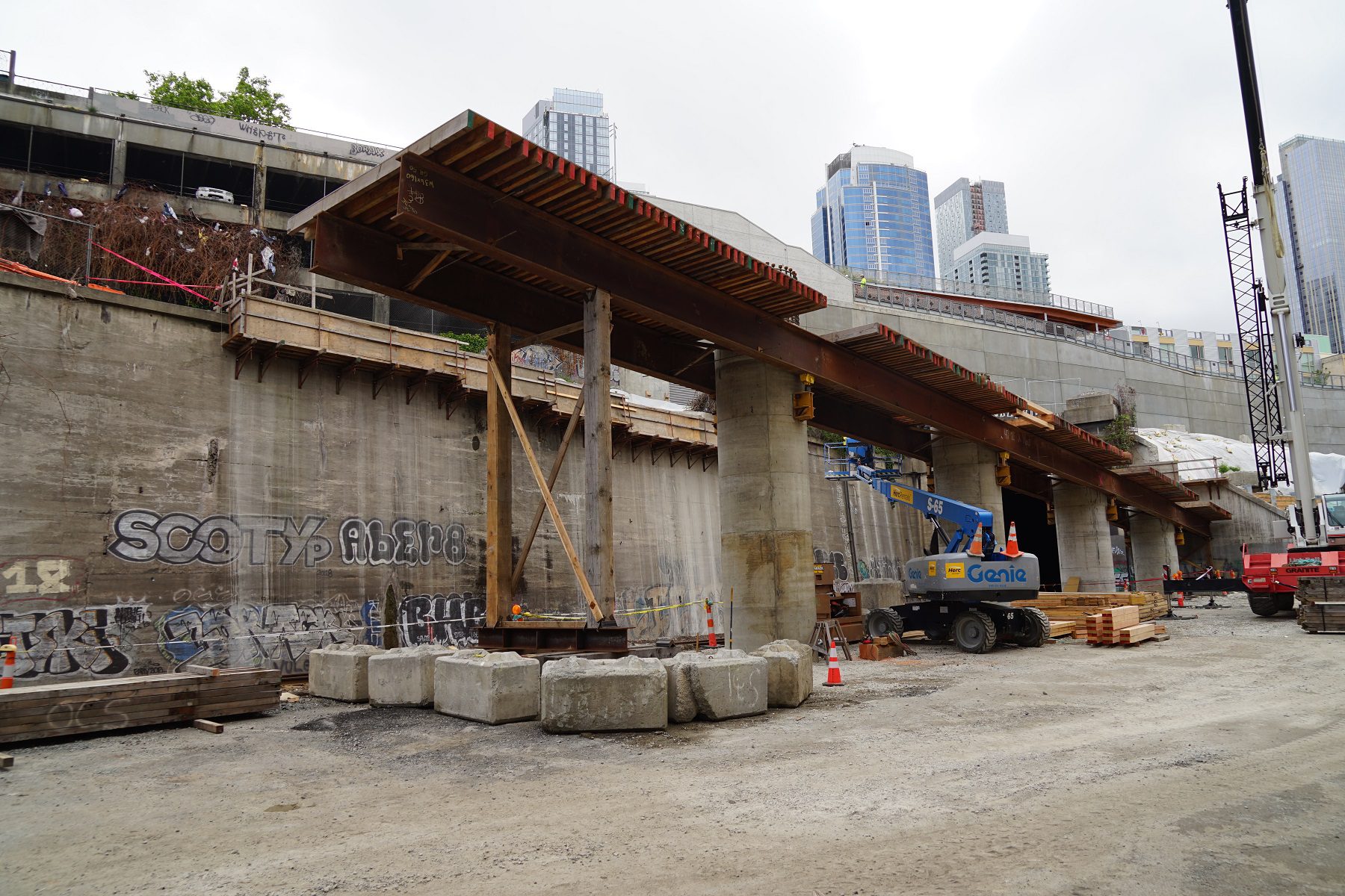 An example of falsework in place for bridge construction. This photo is from construction of the new bridge over the railroad tracks north of Pine St in June 2020. Similar falsework can be expected over Alaskan Way at Marion St. Construction equipment is also present in the middle and right of the image, with large towers in the background.