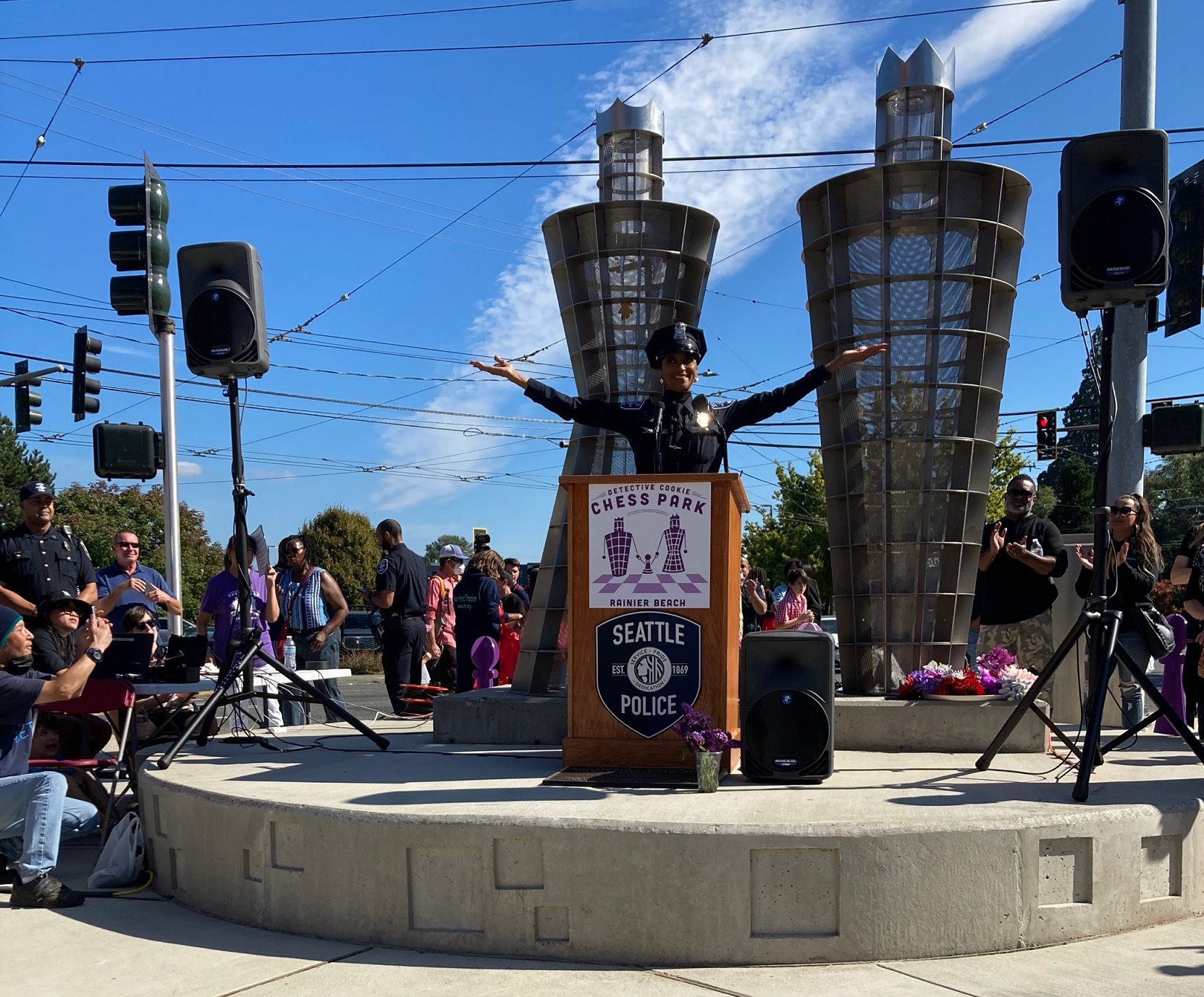 Detective Cookie takes in the moment while speaking at a podium at the newly opened chess park. Detective Cookie stands with her arms raised high in celebration, with other attendees visible in the background. Two large king and queen artistic statues are also in the background.