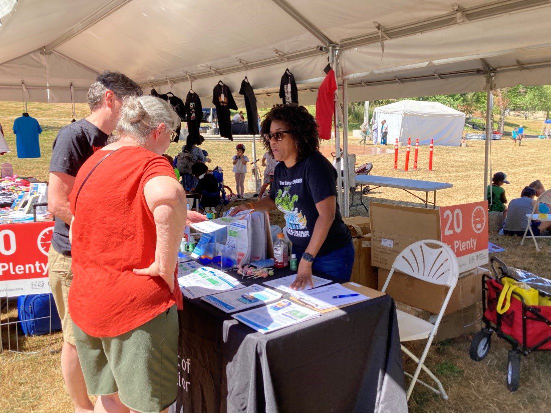 City of Seattle staff member stands behind a SDOT booth in a black graphic tee while speaking to two community members in red and gray shirts about a project happening in the neighborhood. A canopy is above with black, red, and blue shirts hanging off the side.