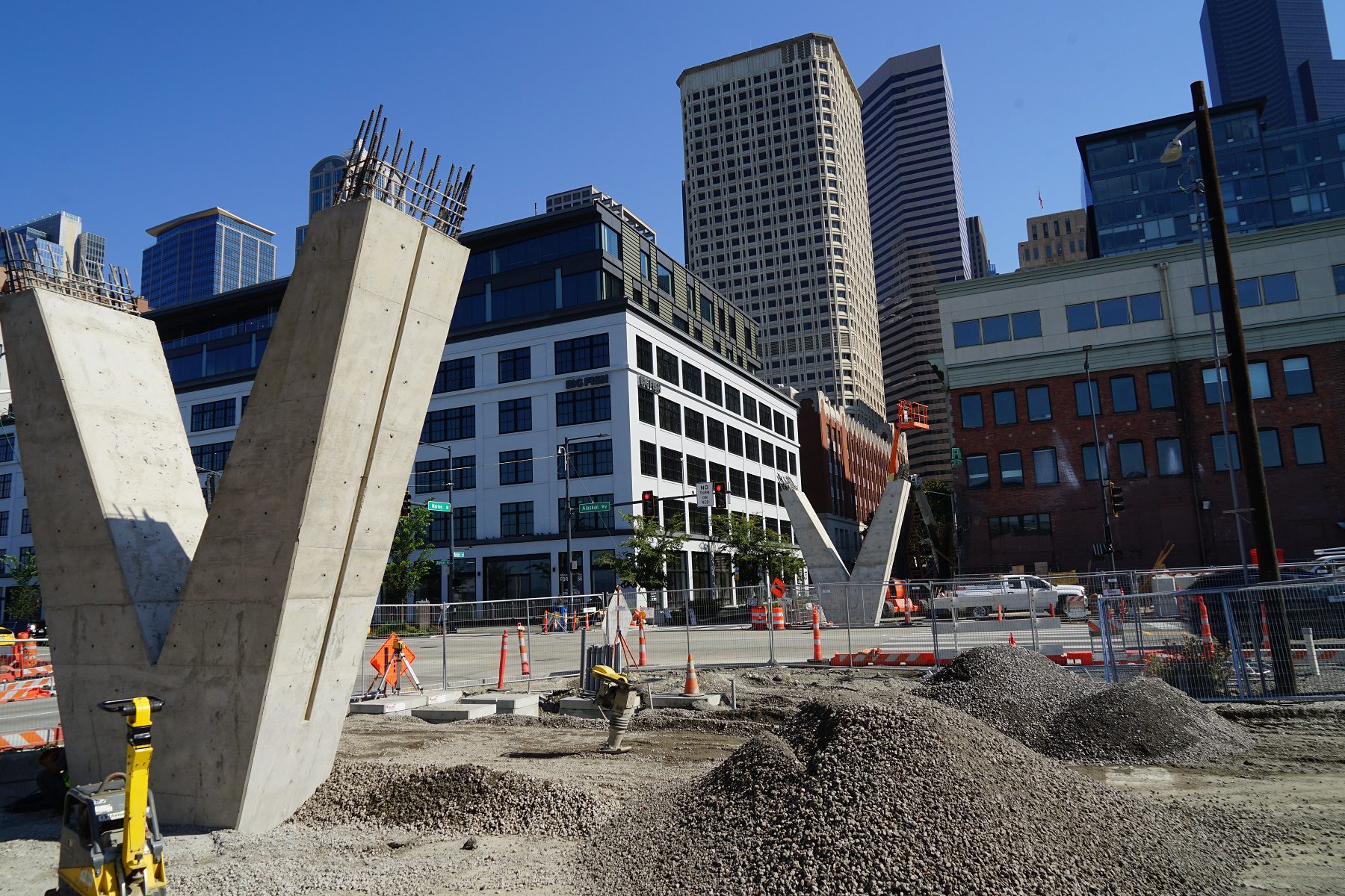 Looking west at the construction site in late August with the new Marion St pedestrian bridge columns in place. Large piles of gravel are in the foreground, with large, tall buildings in the center and background.