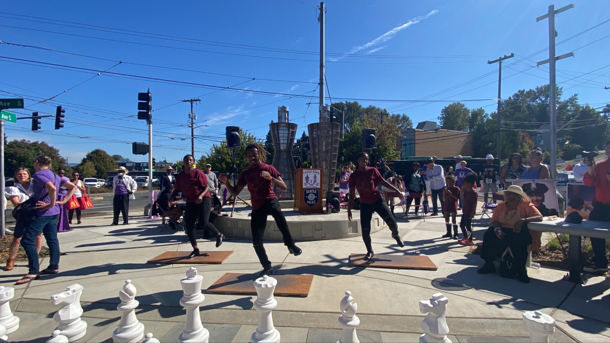 Northwest Tap Connection dancers perform at the new park, as part of the community celebration. Three tap dancers are dancing on wooden boards, with large chess pieces in the foreground, and people watching the performance.