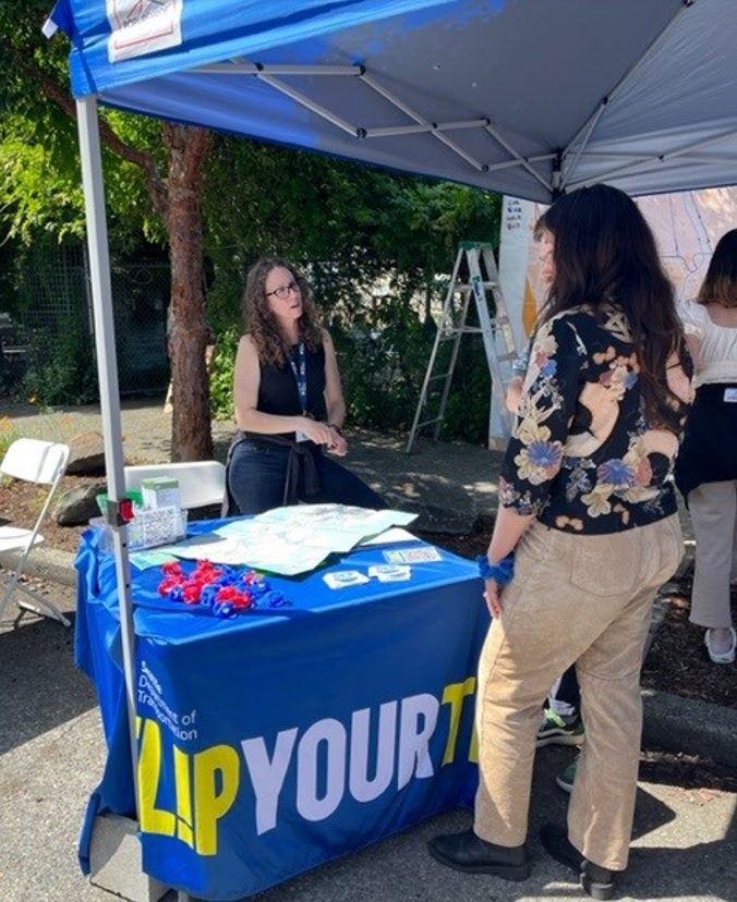 A consultant in a black tank top stands behind a SDOT booth with a Flip Your Trip campaign tablecloth covering the table. SDOT swag is laid out on top of the table as a community member in a black floral shirt speaks to the consultant.  