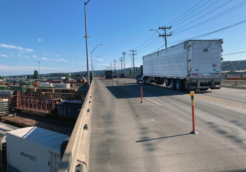 Photo of the 4th Ave S Bridge over the Argo Rail Yard. A large truck carries cargo across the bridge. Several trains below also carrying cargo are on the left side of the photo. Blue skies are above.
