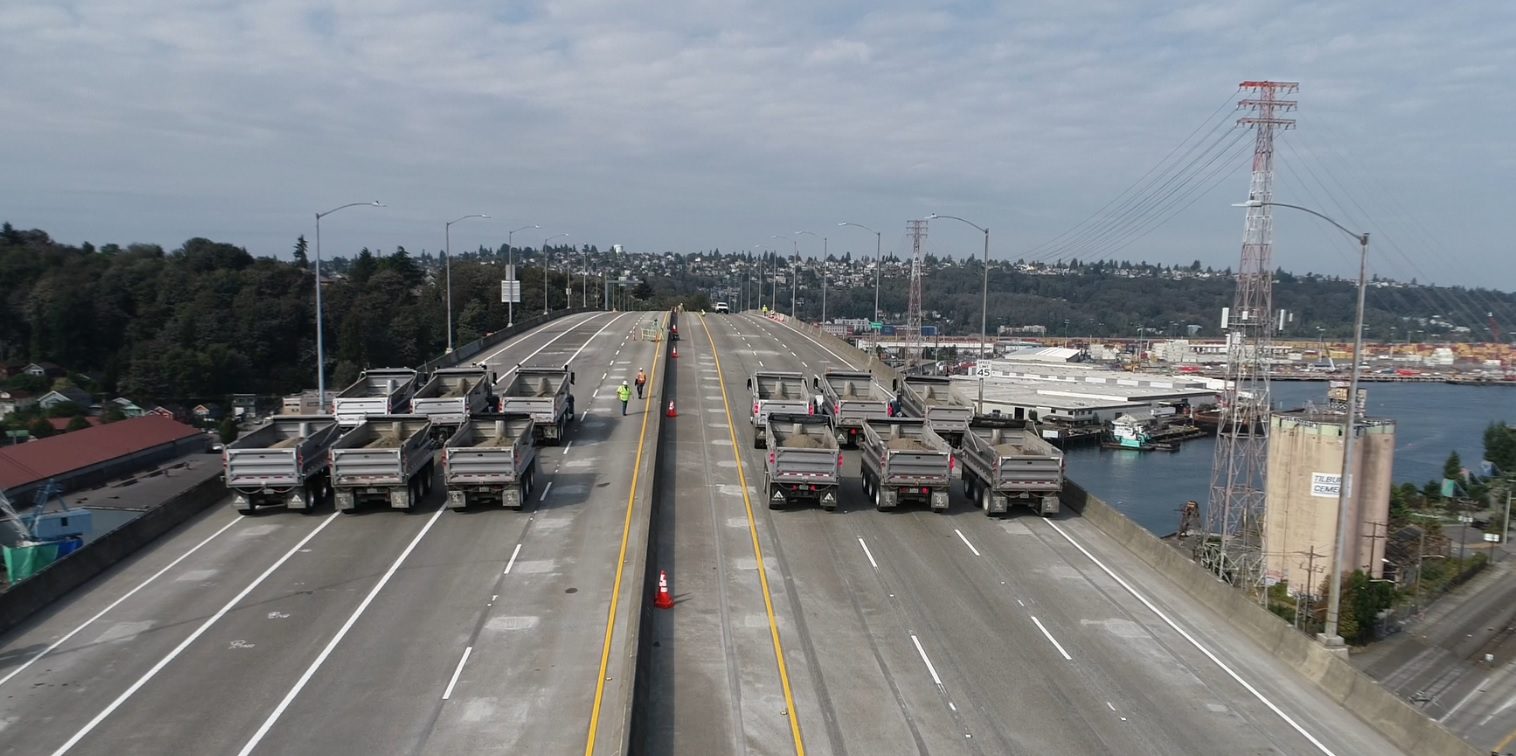 Twelve trucks lined up in preparation for strength testing on the West Seattle Bridge earlier this week. The bridge is in the foreground, with cloudy skies above.