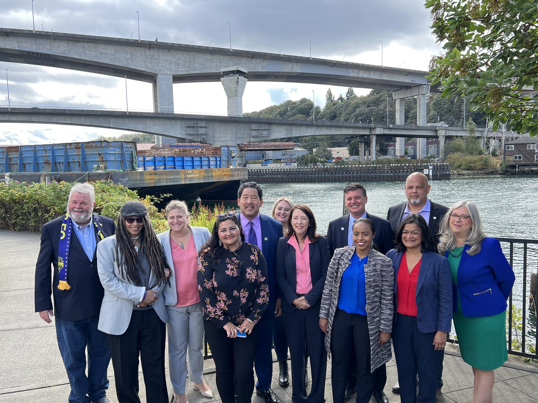 Elected officials, community leaders, City staff members, and other partners stand below the West Seattle Bridge following a press event held on Harbor Island. The high and low bridges are visible, as well as the Duwamish Waterway in the background.