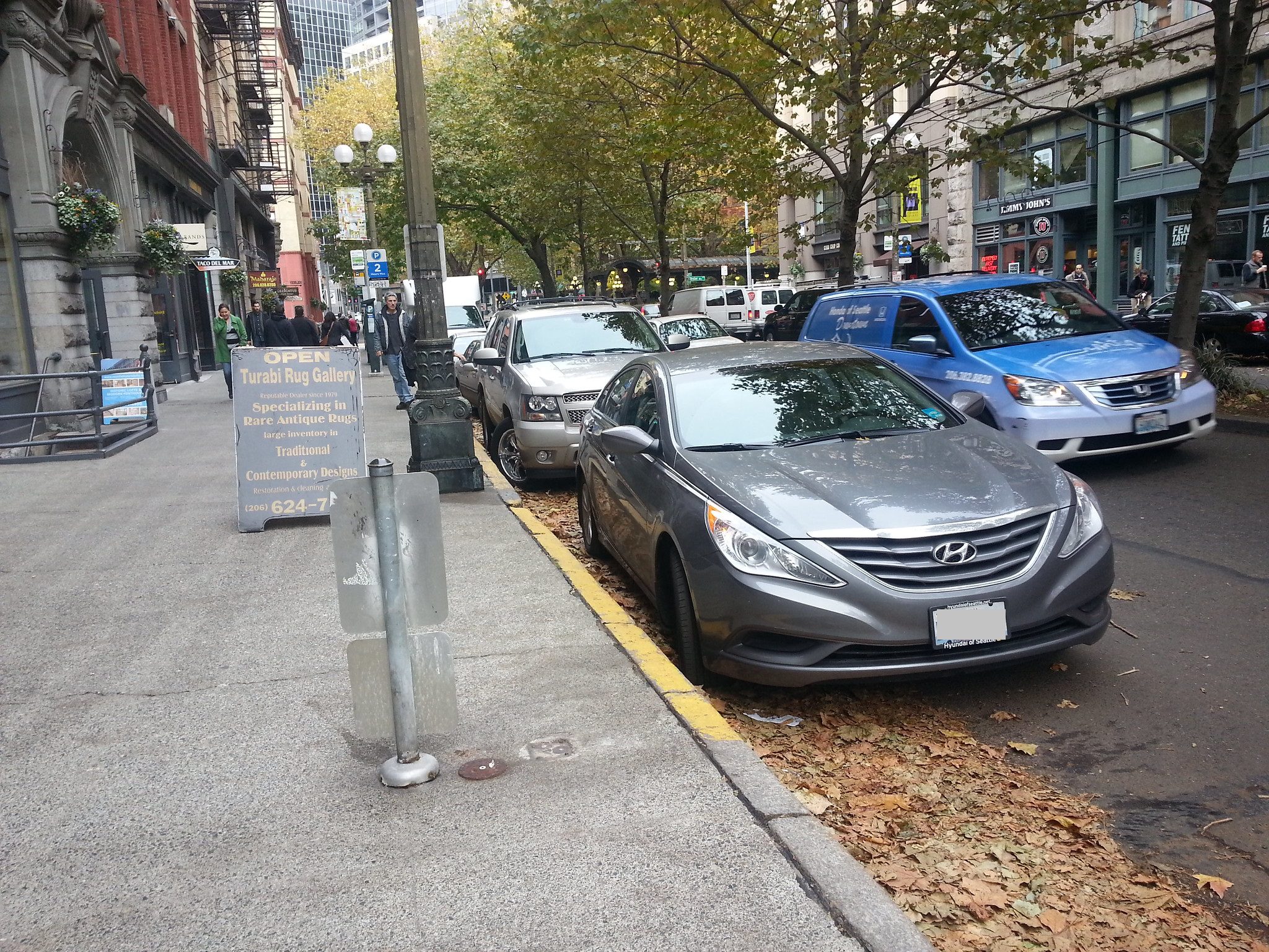 Several parked cars along the curbside in Pioneer Square. Large trees and buildings are in the background, on a fall day.
