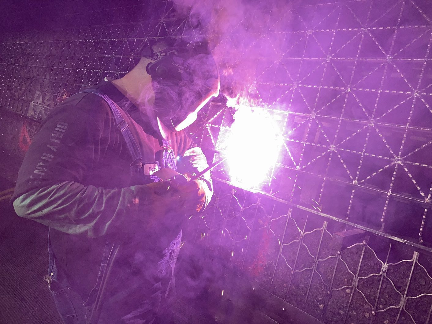 A welder works on the Fremont Bridge deck grating during a recent overnight closure. The person is wearing protective equipment, and the welding creates a bright purple light.