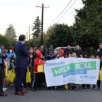 Students begin their walk to school on October 12 in Rainier Beach. Many students hold a sign while walking in a large group, with trees in the background.