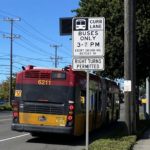 A bus travels in the existing curb lane on 15th Ave W. The red and yellow RapidRide bus is headed away from the camera, and a large white sign notes the hours when the lane is reserved for buses only. A large tree and power lines are also in the background.