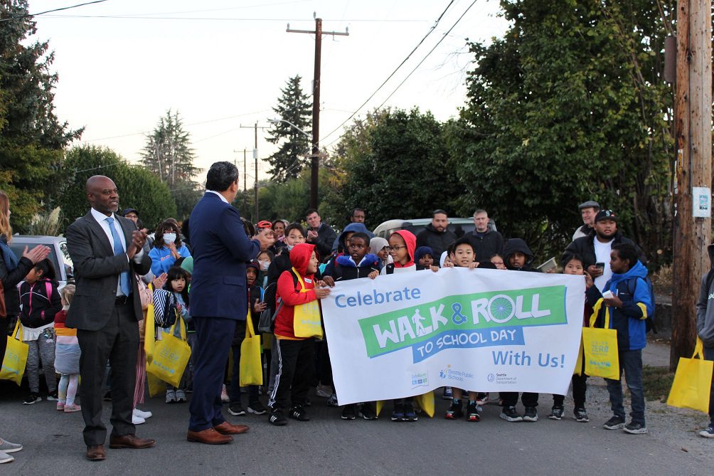 Students begin their walk to school on October 12 in Rainier Beach. Many students hold a sign while walking in a large group, with trees in the background.