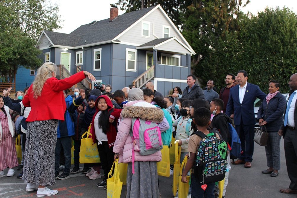 More than a dozen students gather together as they prepare to walk to school. Several adults and City of Seattle officials stand to the right side of the photo. A house and trees are in the background.