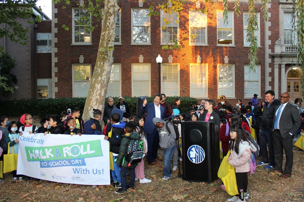 Seattle Mayor Bruce Harrell gives a thumbs up and Nancy Pullen-Seufert shares her remarks while students gather around the podium at Dunlap Elementary School in Rainier Beach. The school's brick building is in the background, as several people look on at the activity.