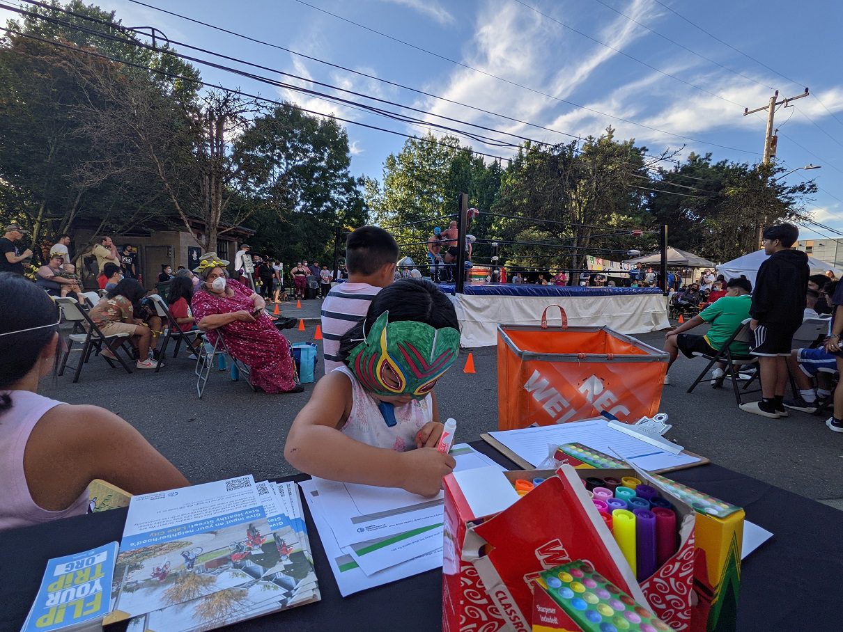 A kid wearing a Lucha Libre mask draws with markers at a recent community event on a Healthy Street in Lake City. Many people sit at the event on a mostly sunny day, with large trees and power lines in the background.