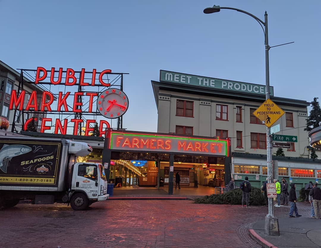 A commercial truck is parked at Pike Place Market in downtown Seattle. People stand in the right side of the photo near signs, buildings, and the brick street.